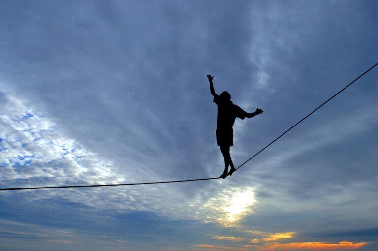 Silhouette of young man balancing on slackline, sun and clouds behind. Slackliner balancing on tightrope between two rocks, highline silhouette.