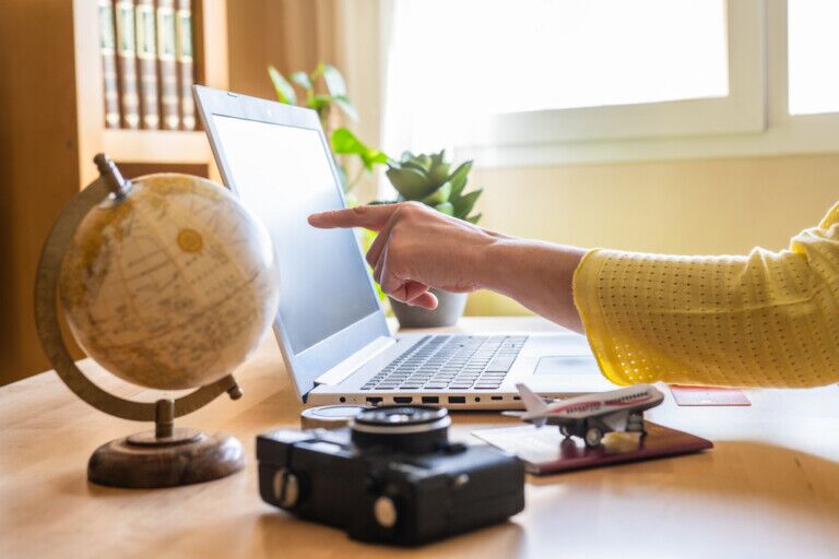 AI search Woman pointing at laptop screen while planning a trip on a booking website, surrounded by a globe, camera, passport, and airplane model on the desk, creating an organized itinerary